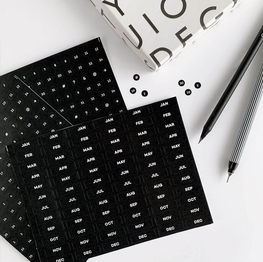 Black HELLO TIME planner stickers with months, numbers, and symbols on white desk
