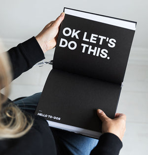 Person holding HELLO TIME planner open to black page with bold white text “OK LET’S DO THIS” and “HELLO TO-DOs” section title