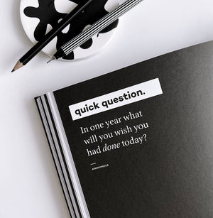 HELLO TIME planner open to black page with white quote “In one year what will you wish you had done today?” and pencils on abstract coaster