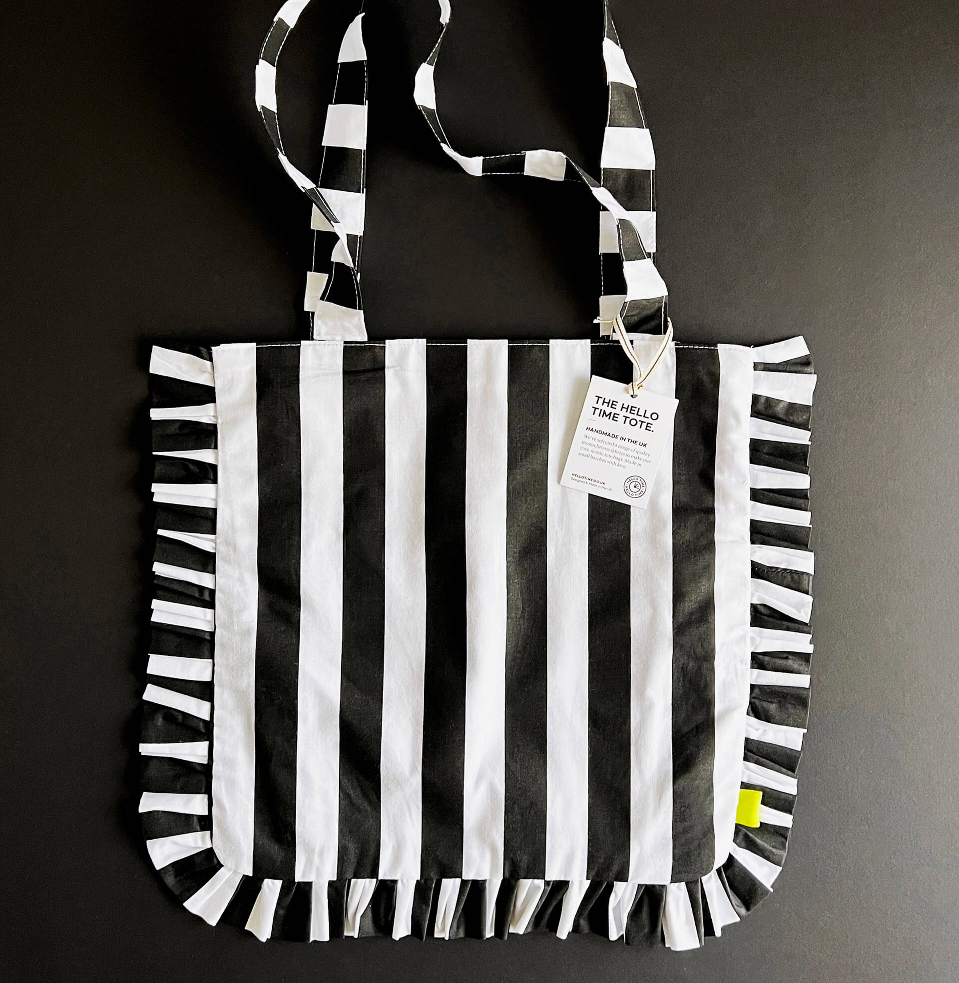Black and white striped HELLO TIME tote bag with ruffled edges, laid flat with branded tag attached