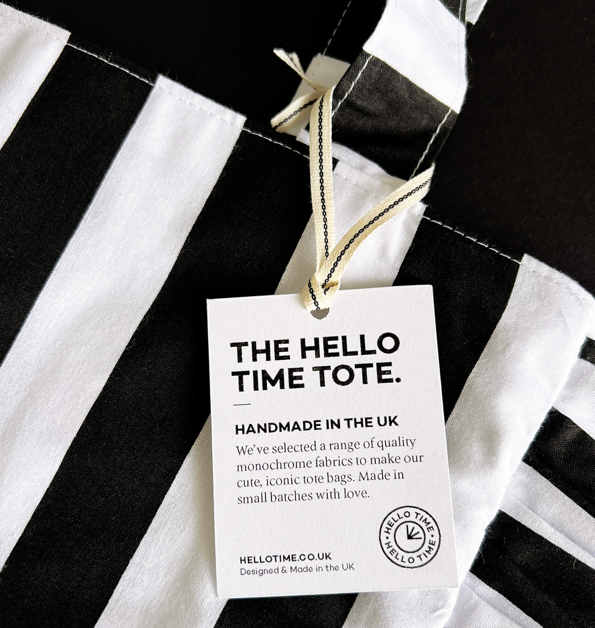 Close-up of HELLO TIME tote bag tag on black and white striped fabric, highlighting handmade UK label