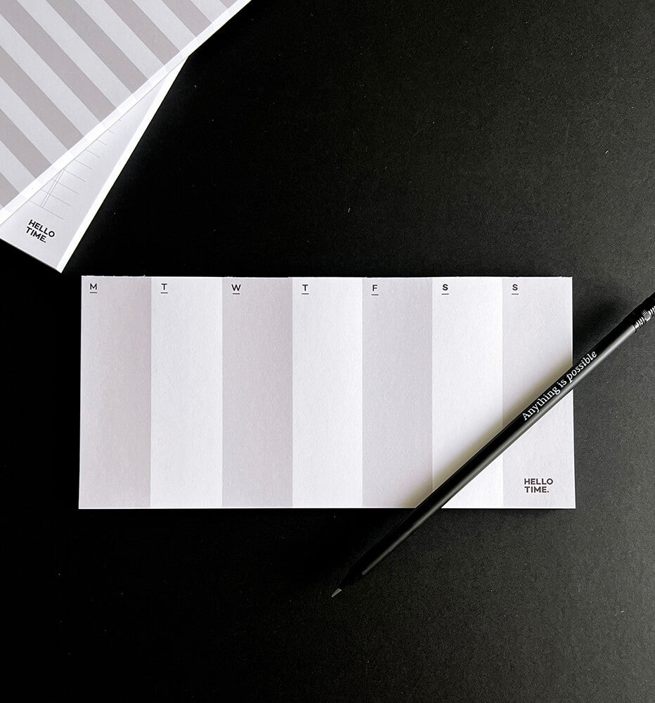 DL-sized white weekly desk pad with minimal layout and pencil, showing Monday to Sunday columns on black background.