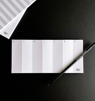 DL-sized white weekly desk pad with minimal layout and pencil, showing Monday to Sunday columns on black background.