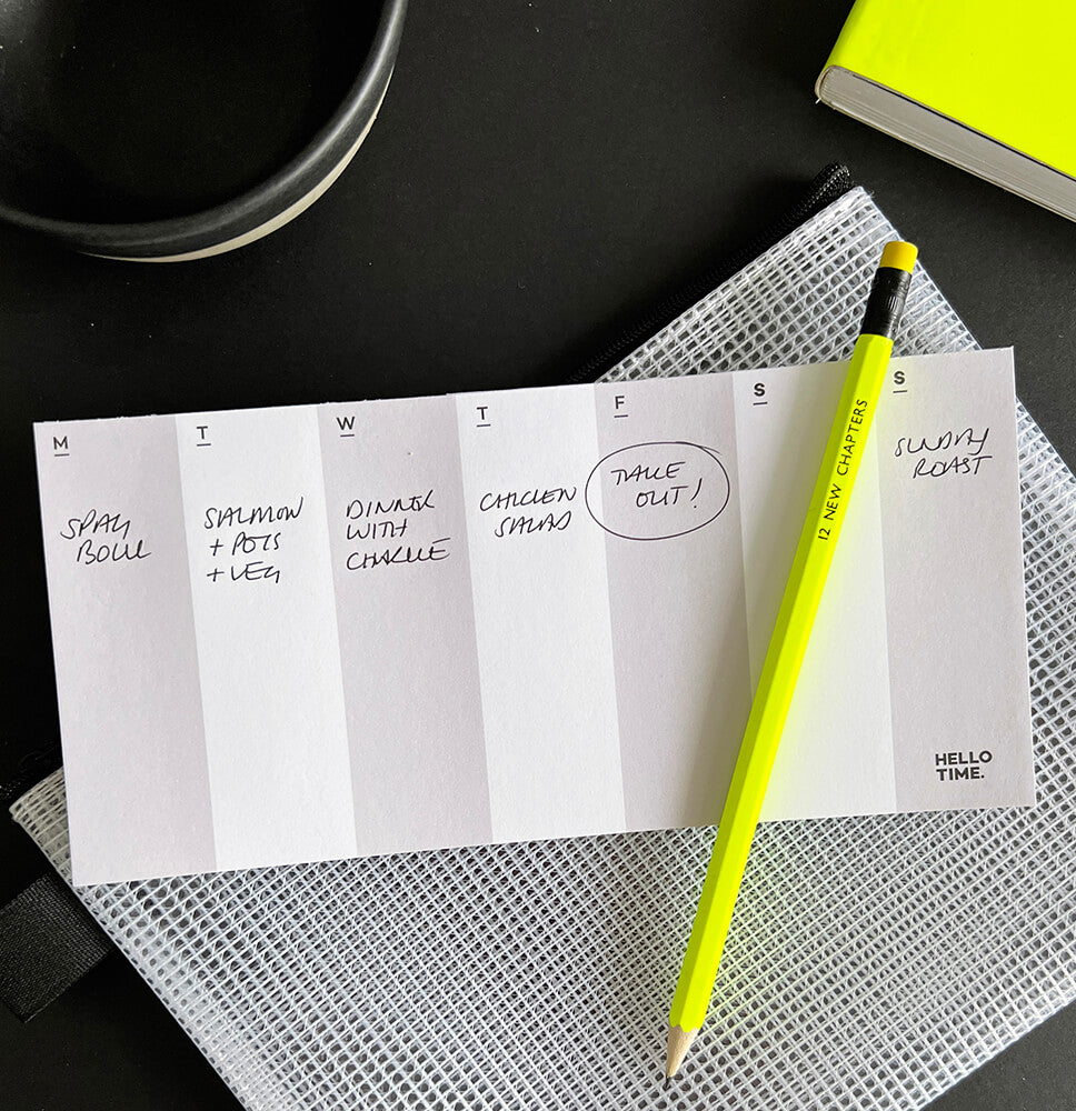 DL-sized weekly pad filled with handwritten meal plan, neon yellow pencil and mesh pouch on black background.