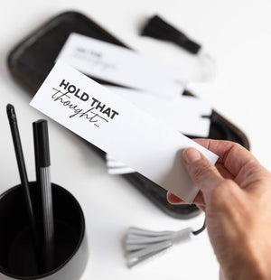 Hand holds “HOLD THAT THOUGHT…” bookmark over an open book with white leather tassel