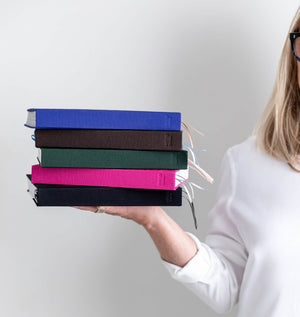 Woman holding a stack of five HELLO TIME planners in blue, brown, green, pink and black with ribbon bookmarks showing.