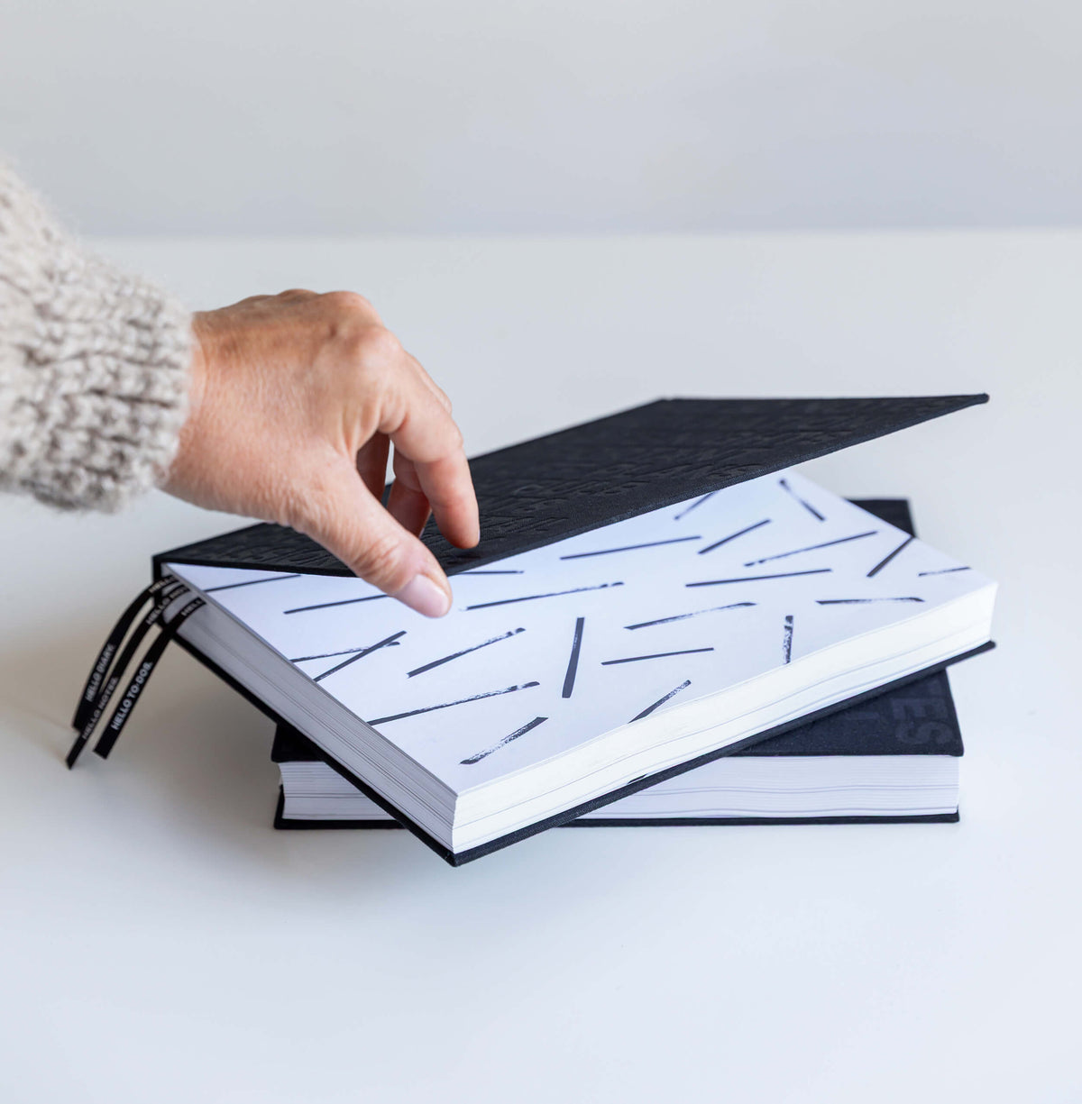 Hand lifting cover of black HELLO TIME planner to reveal black line-pattern endpaper, black ribbons, second planner below.