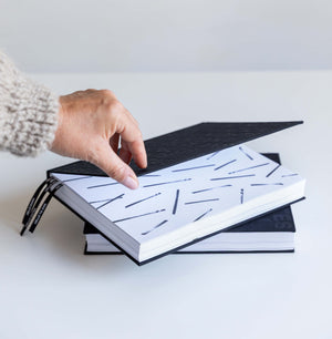 Hand lifting cover of black HELLO TIME planner to reveal black line-pattern endpaper, black ribbons, second planner below.