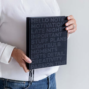 Person in white shirt holding black HELLO TIME planner with embossed cover text and black branded ribbon bookmarks.