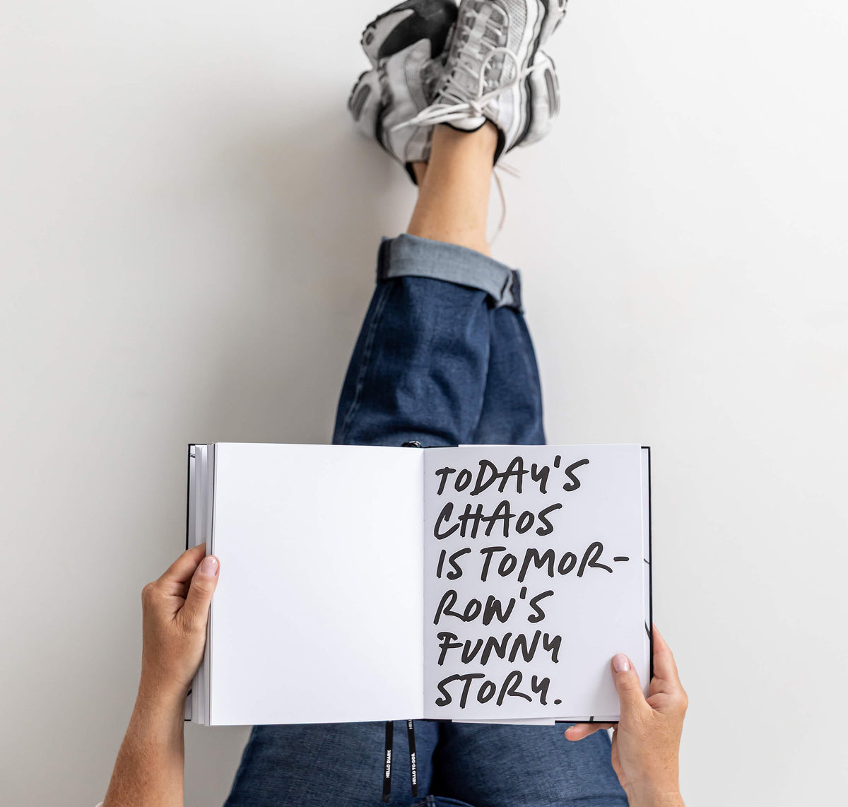 Person lying back with feet up a wall, holding open HELLO TIME planner with handwritten “Today’s chaos is tomorrow’s funny story”.