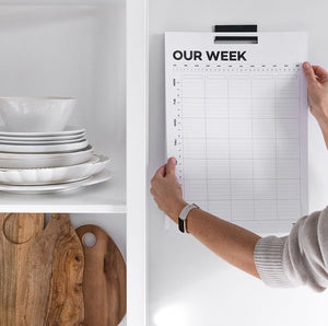 Person placing Hello Time weekly family wall planner on kitchen wall near open shelf with dishes and boards