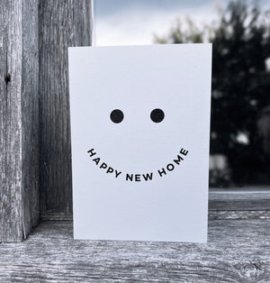 White card with black smiley face design and curved text “Happy New Home,” displayed against wood and sky.