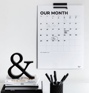 Black and white monthly wall planner with handwritten dates, hanging on a white wall