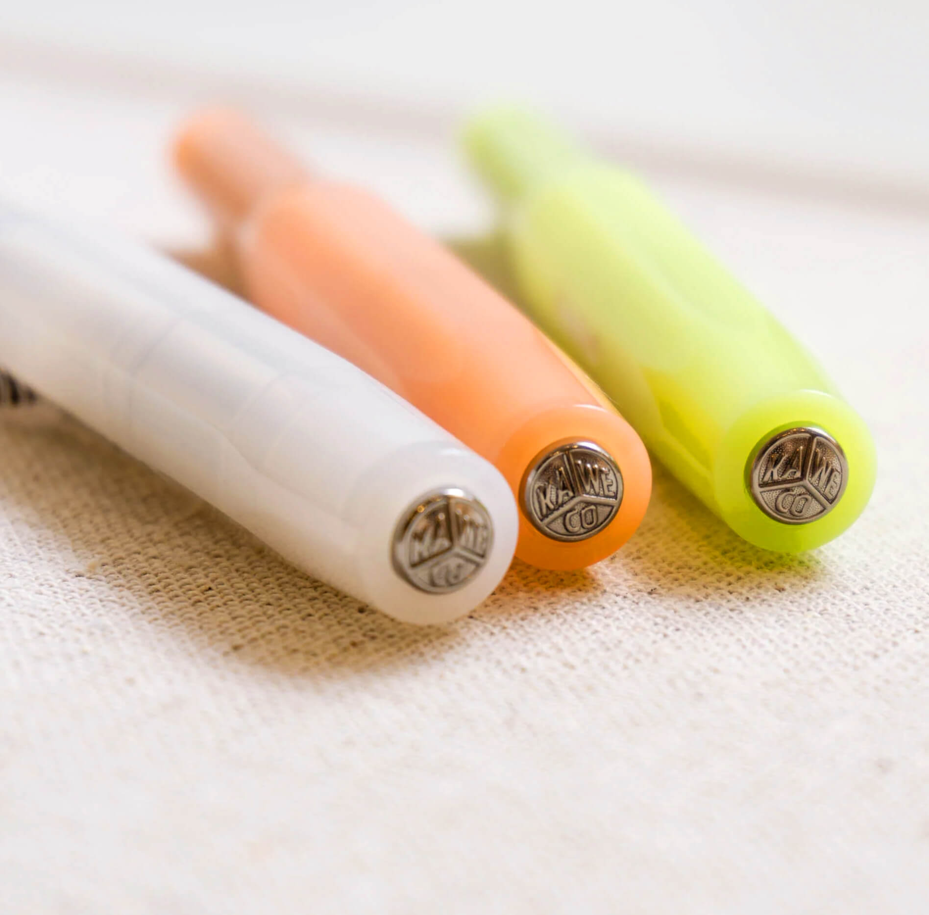 Three Kaweco Sport fountain pens in white, orange, and lime with chrome logo tops, shown angled on a fabric surface