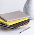 HELLO TIME Luxe Leather Notebook Covers in clay, yellow and black stacked on wooden tray with stripy Edding pen nearby
