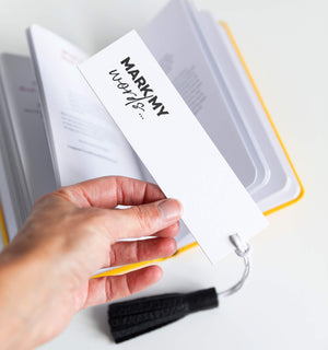 Hand holding black leather tassel bookmark in use with “MARK MY WORDS…” on it