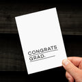 Hand holding a minimal white graduation card with bold Congrats Grad text.