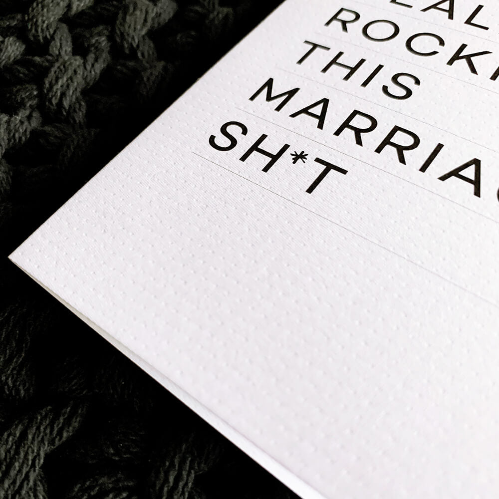 Close-up of white textured card with bold black text "We’re really rocking this marriage sh*t."