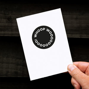 White congratulations card with bold black Whoop Woo design, held in hand.