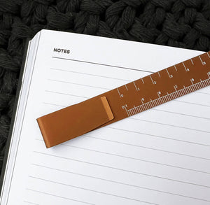 Copper ruler bookmark on lined notebook page, showing clip and measuring scale.