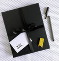 Wrapped black gift box with black ribbon, gift card, yellow marker, and black striped pen