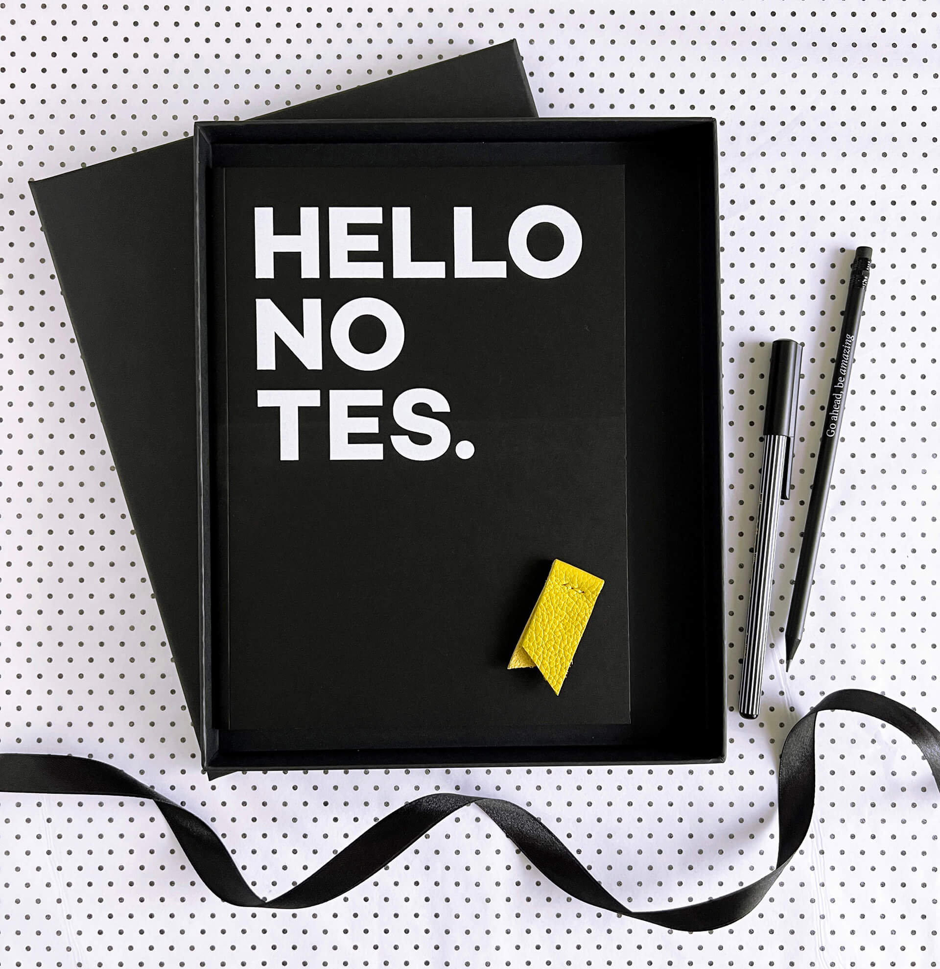 Open black gift box with HELLO NOTES notebook, yellow leather marker, pencil and pen on spotty paper