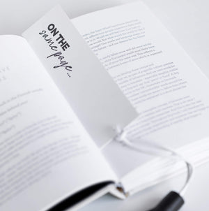 White card bookmark with black leather tassel saying “ON THE SAME PAGE…” inside an open book