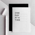 White greetings card with bold black text reading One Day At A Time, black envelope behind.