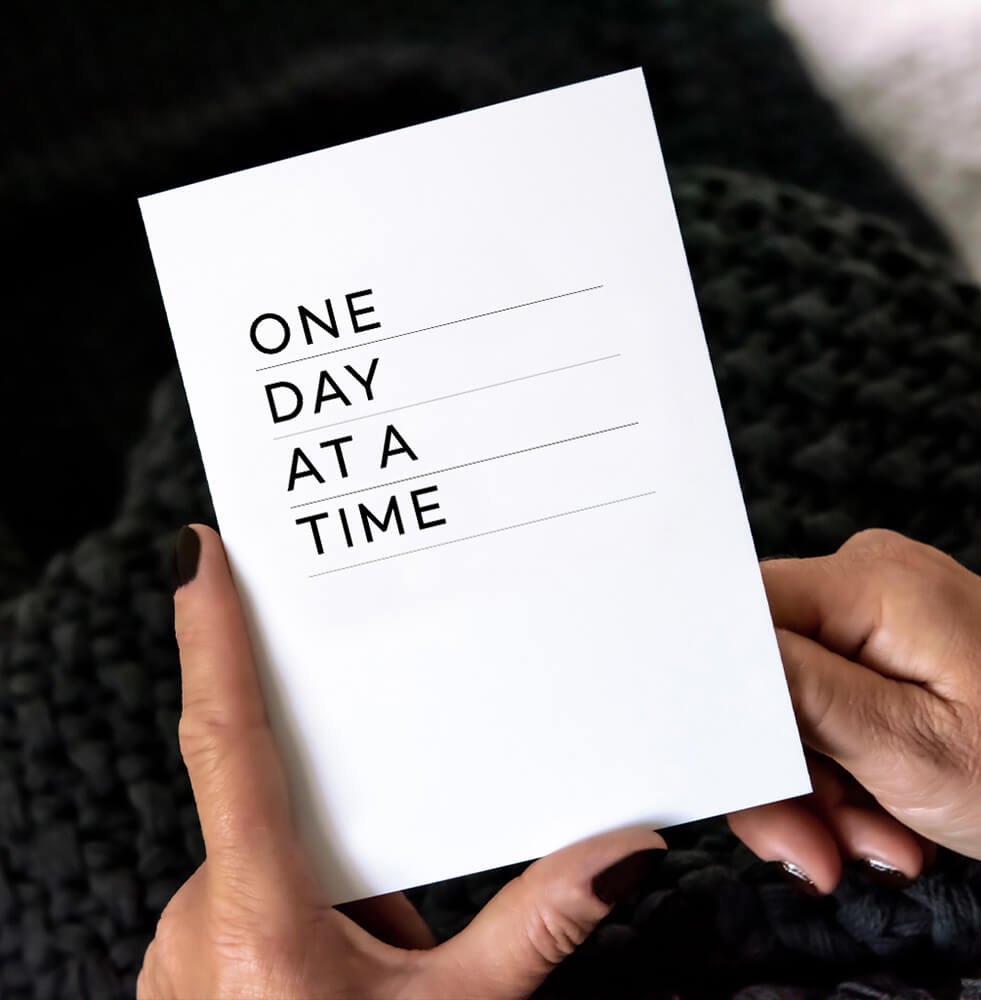 White greetings card with bold black text reading One Day At A Time, black envelope behind.