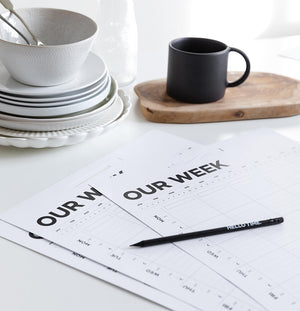 Hello Time weekly family planner sheets on white kitchen table with pencil, cup, and stacked crockery nearby