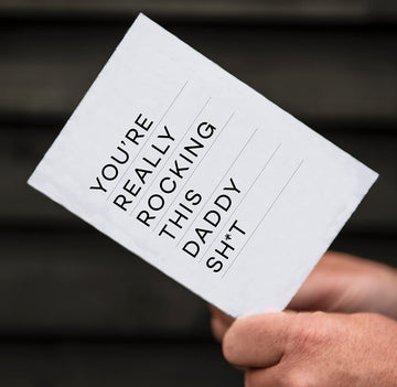 White card with bold black text reading "You’re really rocking this daddy sh*t" held in hand.