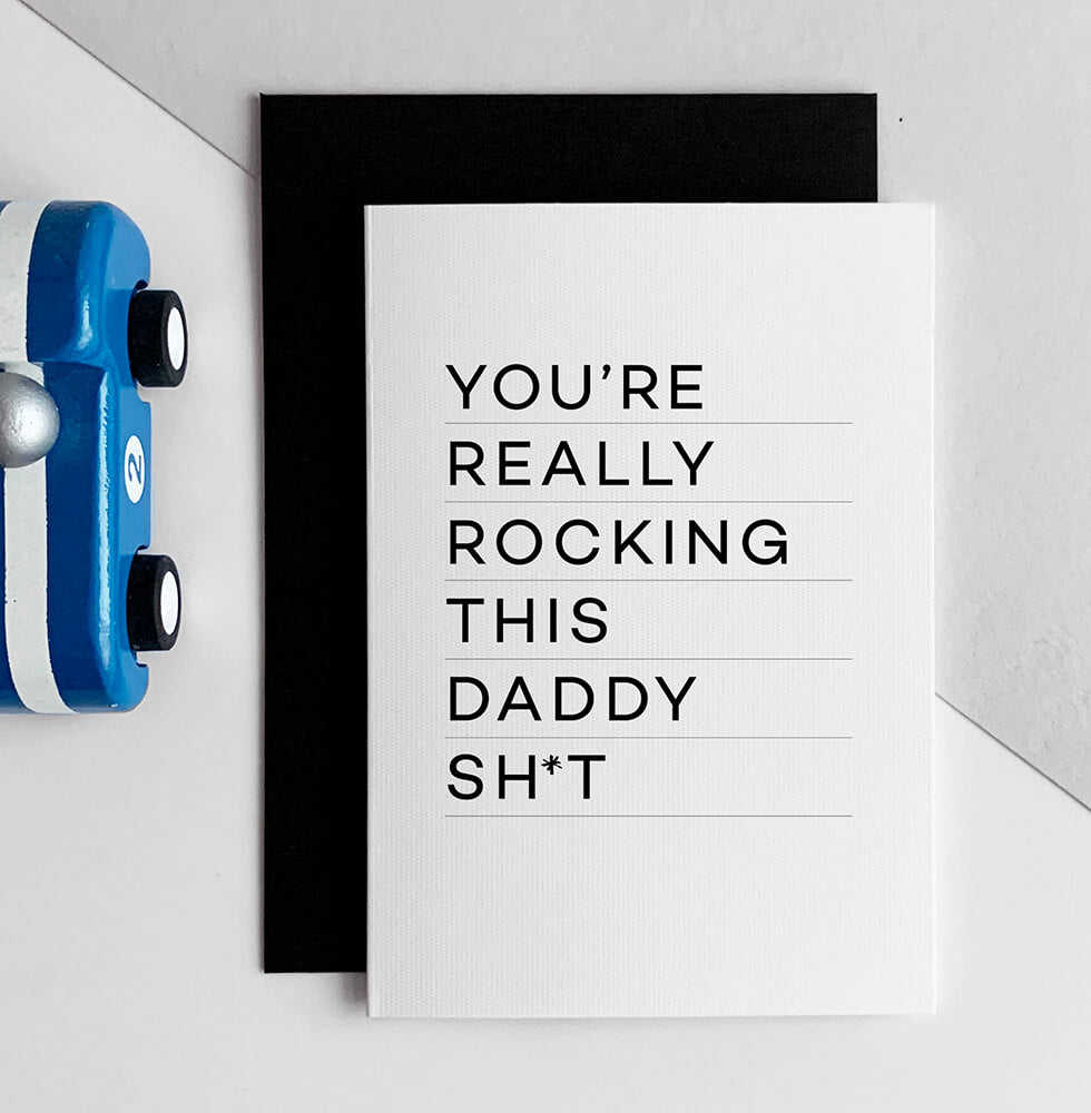 White card with bold black text reading "You’re really rocking this daddy sh*t" held in hand.