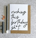 White birthday card with black handwritten text reading ‘rocking this birthday sh*t’, paired with a kraft envelope.