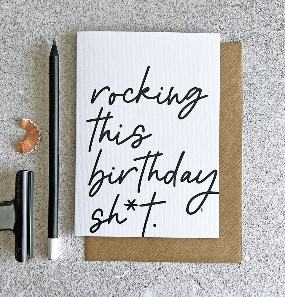 White birthday card with black handwritten text reading ‘rocking this birthday sh*t’, paired with a kraft envelope.