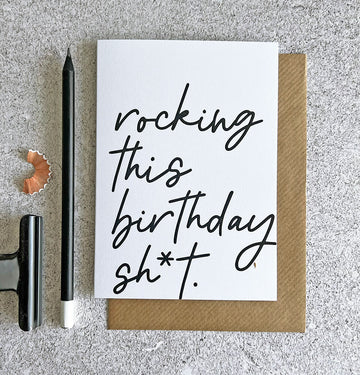 White birthday card with black handwritten text reading ‘rocking this birthday sh*t’, paired with a kraft envelope.