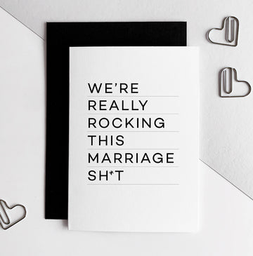 White anniversary card with bold black text reading "We’re really rocking this marriage sh*t," black envelope behind.