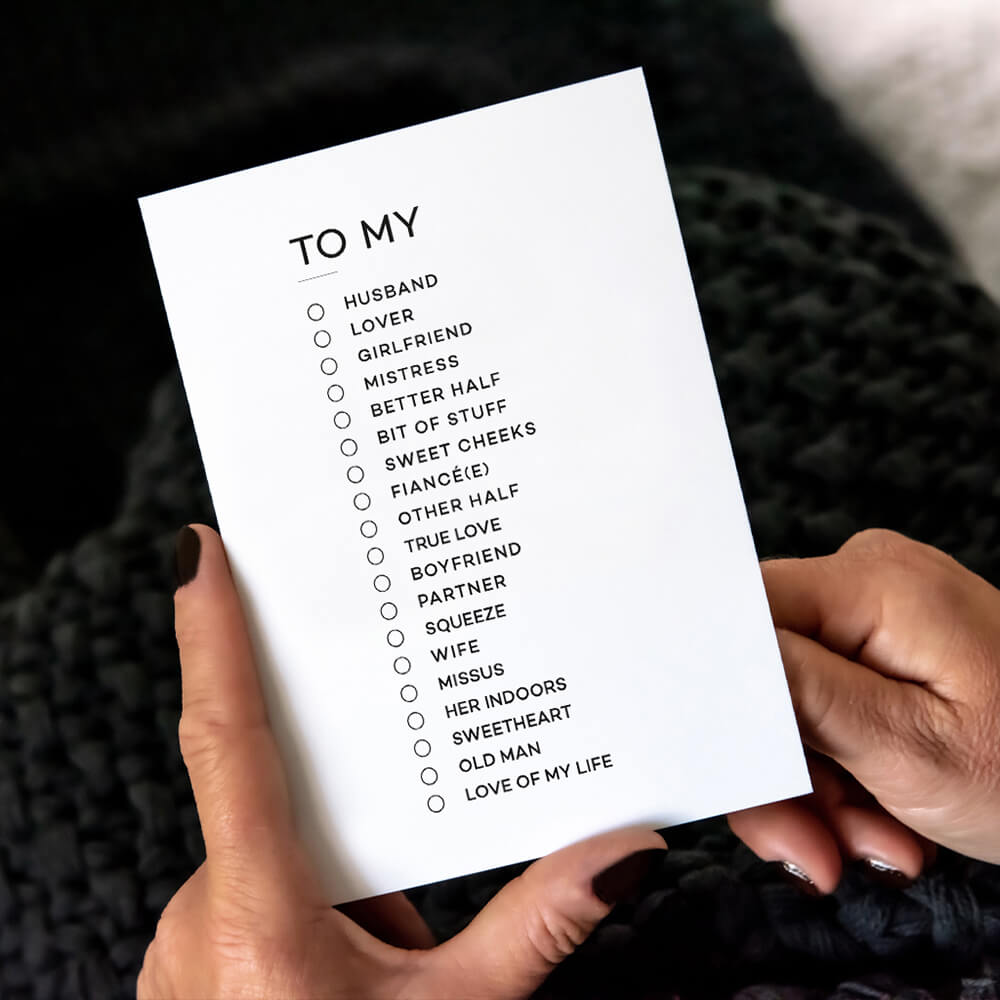 Person holding white anniversary card with "To My" partner checklist, black background.