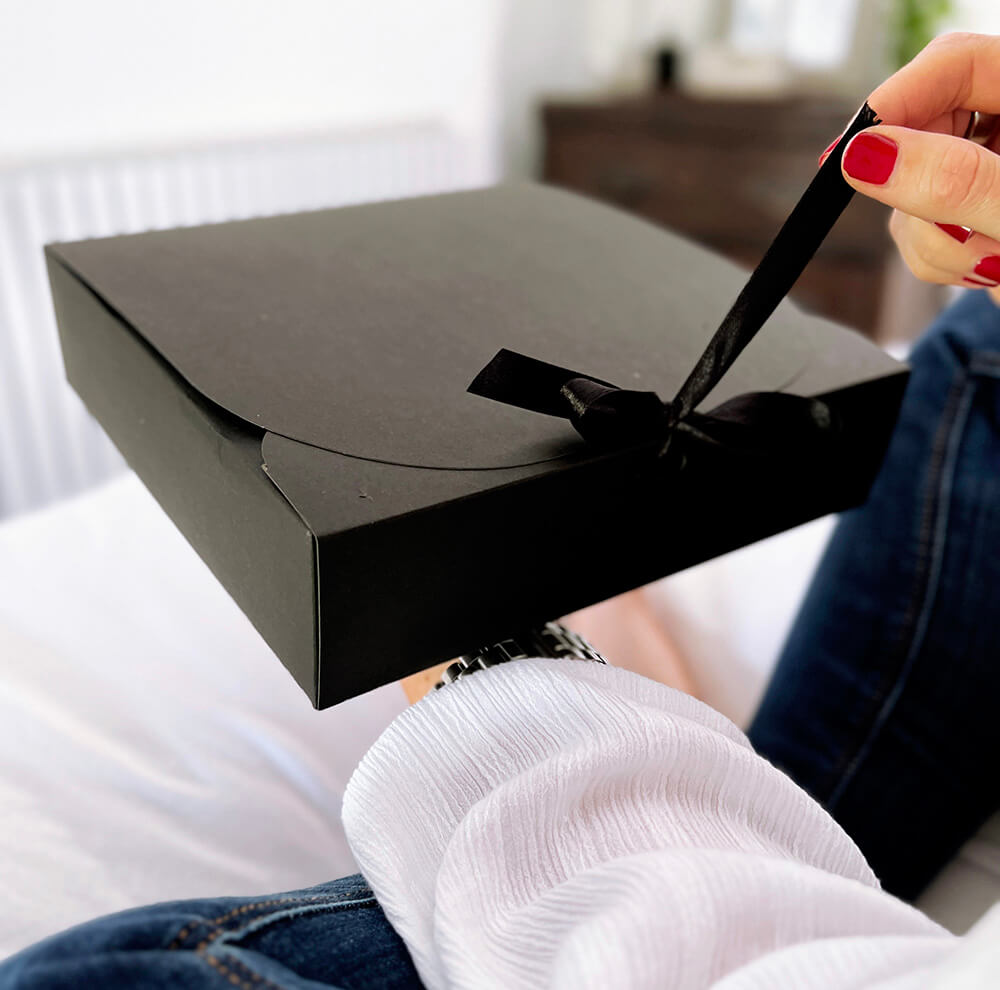 Person in white blouse and jeans untying black ribbon on HELLO TIME gift box with black packaging