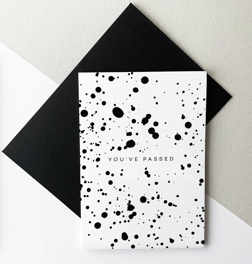 Black and white greeting card with splatter design and text “You’ve Passed,” paired with black envelope.